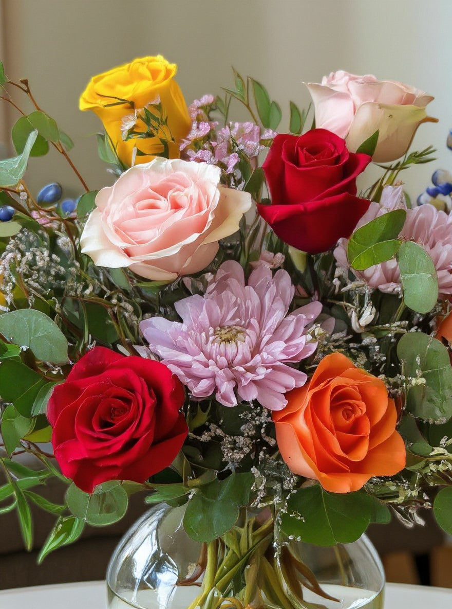 Lakewood Florist Happy Blooms arrangement: vibrant red, pink, orange, and yellow roses with purple mums and blue accents in a clear glass vase.